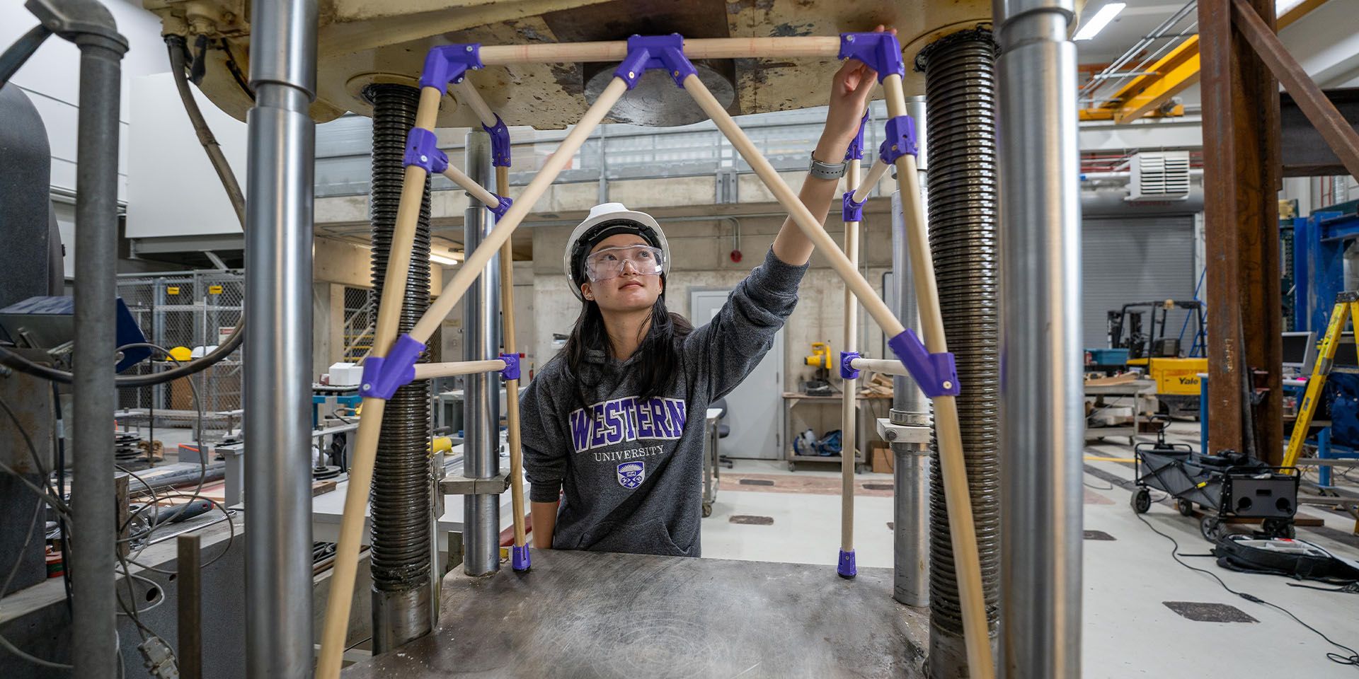 A student wearing a grey Western hoodie with safety goggles and a hard hat is inspecting a structure inside a lab